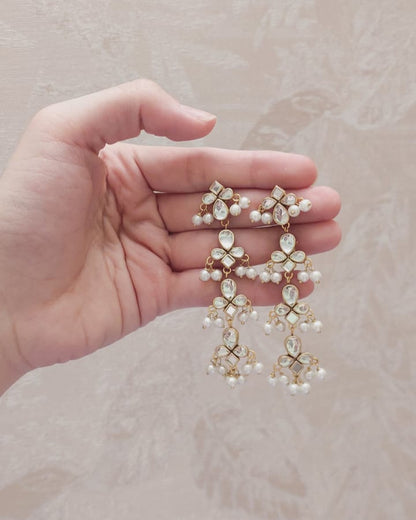Hand holding a pair of floral earrings with pearl-like stones against a beige background
